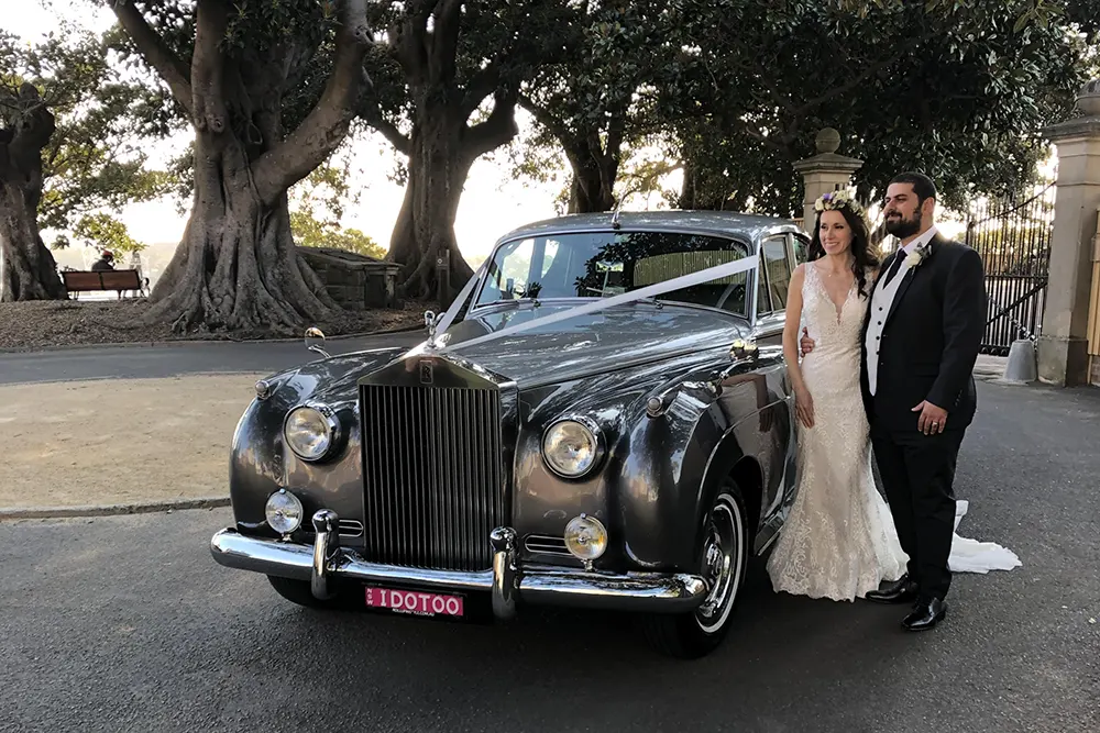 a bride in white dress and groom in black suit next to a classic rolls royce wedding car